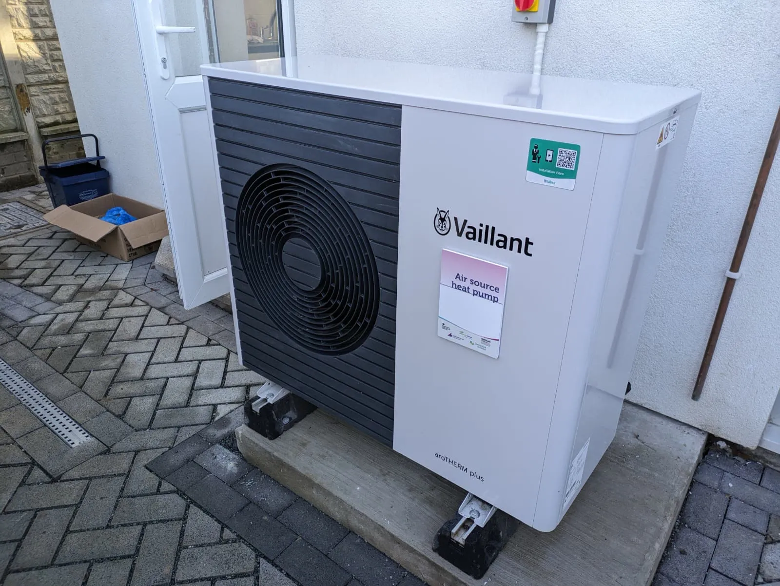 Unité extérieure d'une pompe à chaleur air-eau installée au pied d'une maison, sur un pavage
